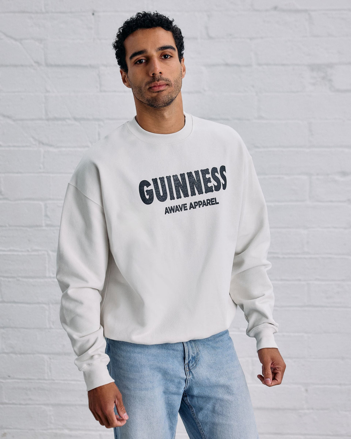 Man wearing a white 'Guinness Apparel' sweatshirt against a white brick wall.