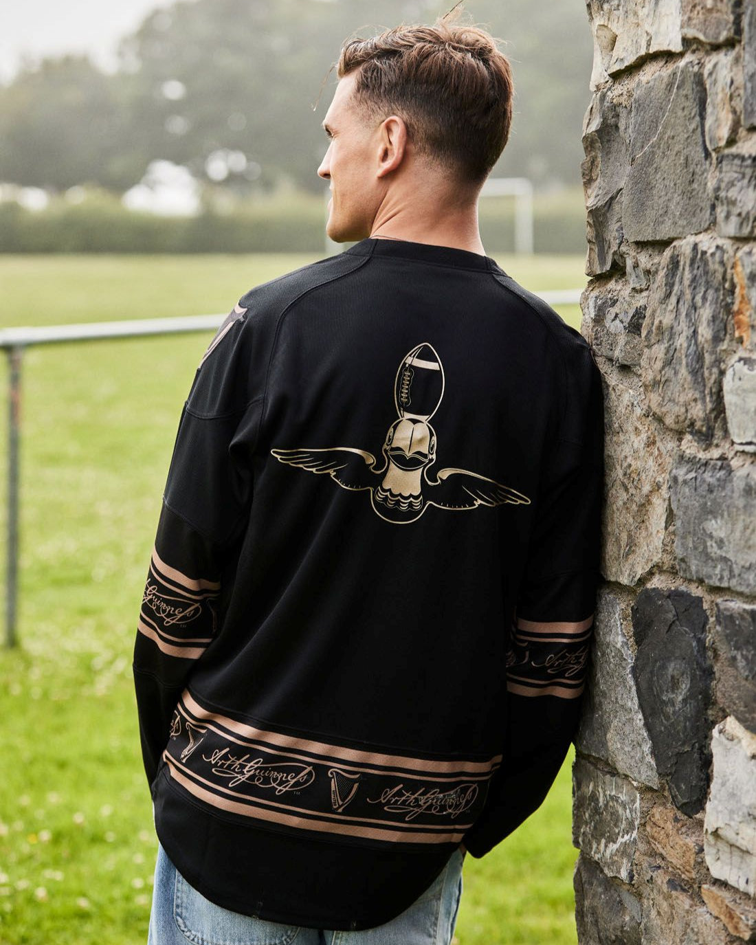 Man wearing a black jacket with a gold design, leaning against a stone wall outdoors.