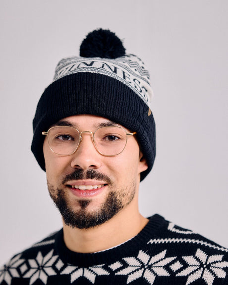 Man wearing a black knit beanie with a pom-pom and a black and white patterned sweater against a gray background