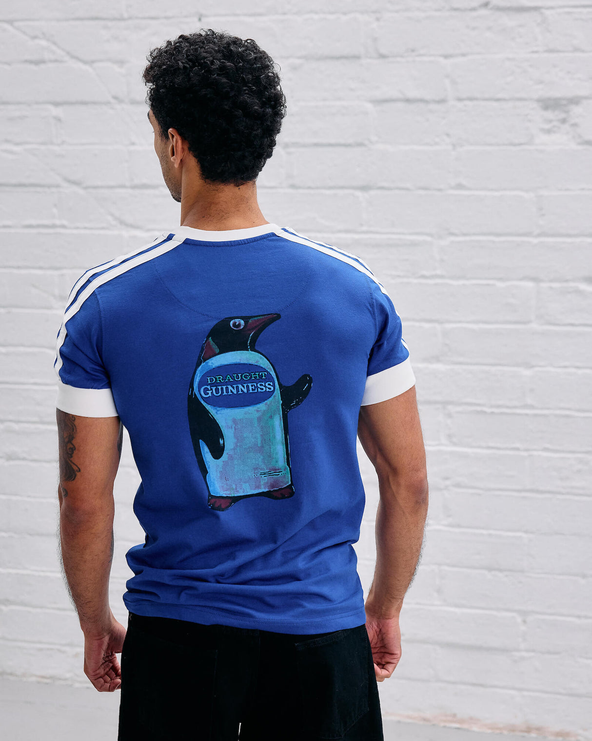 Person wearing a blue t-shirt with a Guinness penguin logo on the back against a white brick wall.