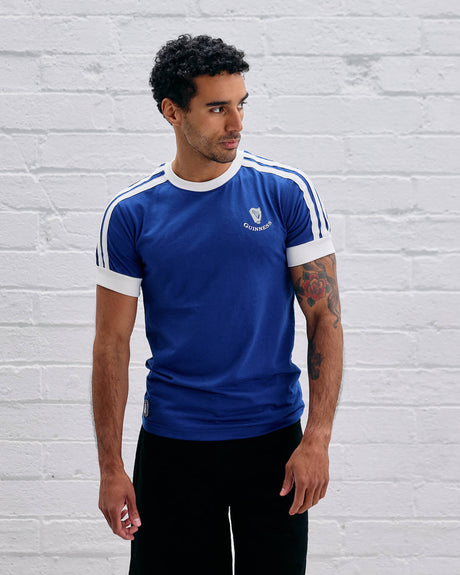 Man wearing a Guinness blue t-shirt with white stripes against a light gray brick wall