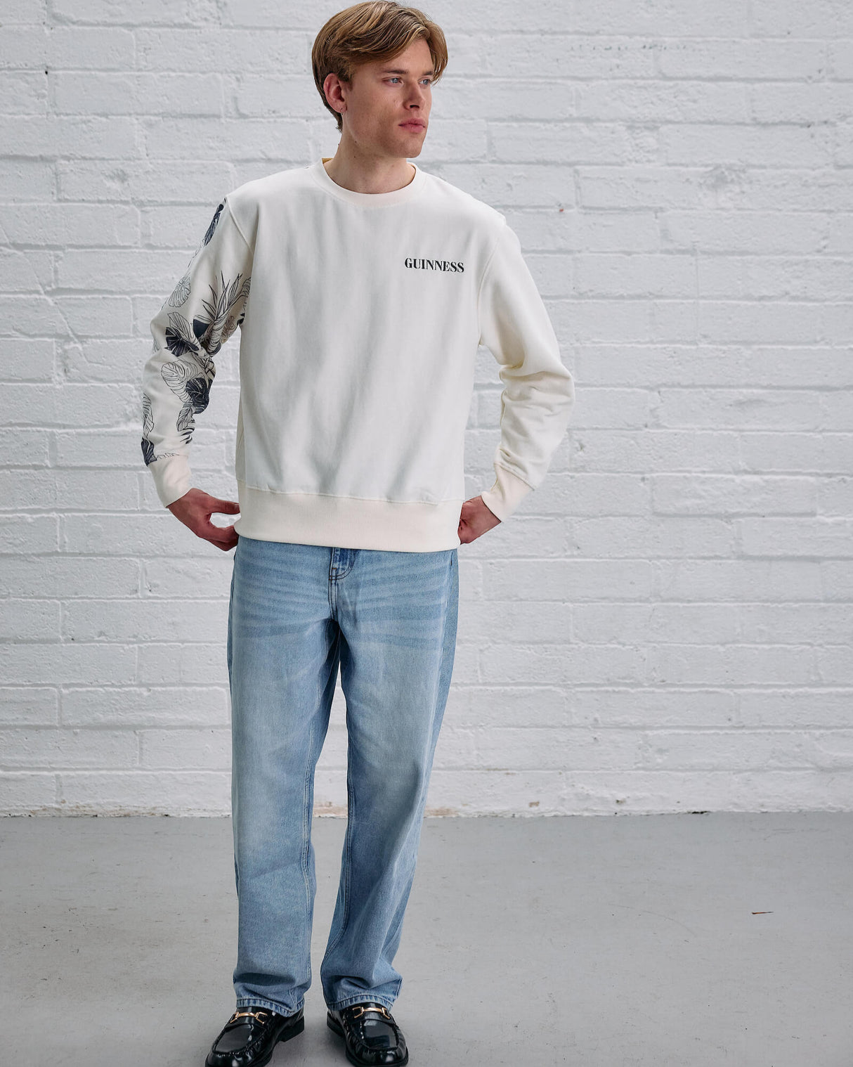 Person wearing a white sweatshirt with brand logo and light blue jeans against a white brick wall.