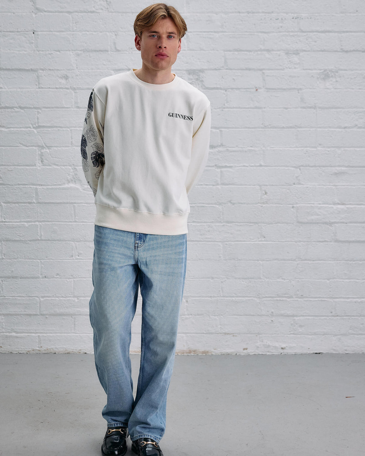 Person wearing a white sweatshirt and blue jeans standing against a white brick wall.