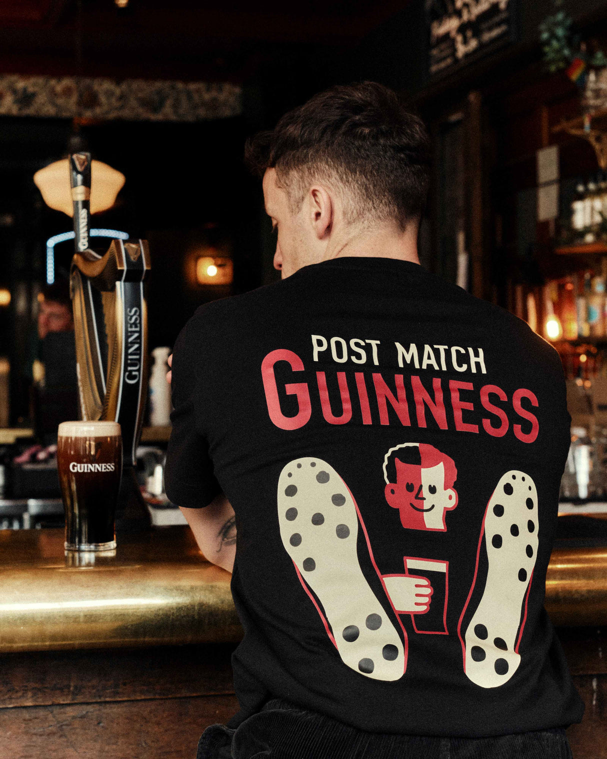 Person wearing a black t-shirt with 'Post Match Guinness' text and graphics in a bar setting.
