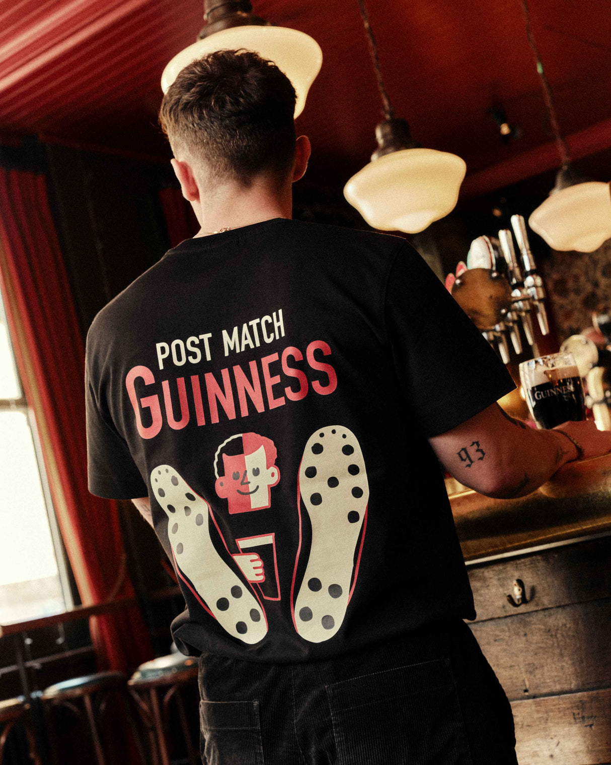 Person wearing a black t-shirt with 'Post Match Guinness' design in a bar setting