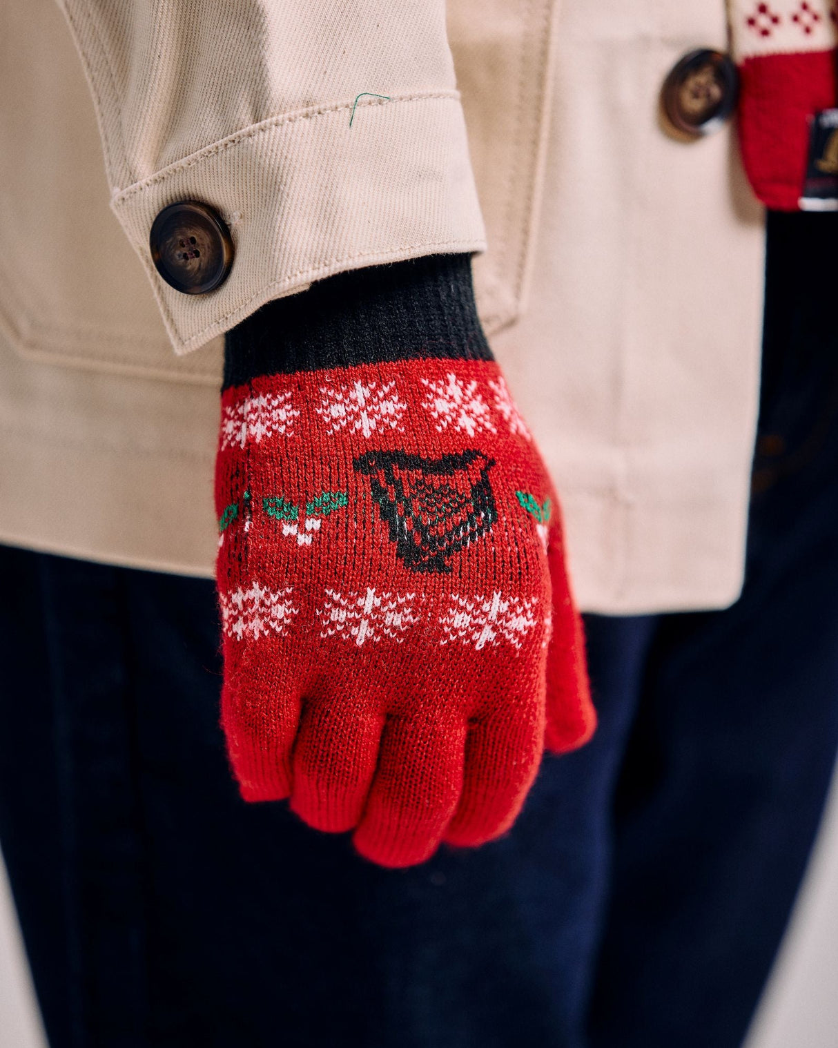 Red knitted glove with snowflake pattern held by a person wearing a beige coat.
