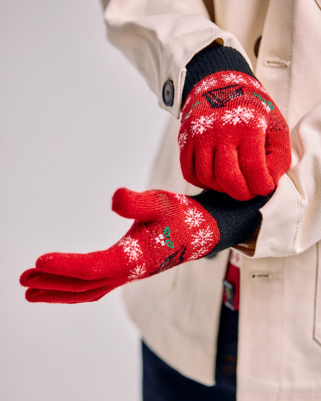 Red gloves with snowflake patterns worn by a person in a beige coat against a light gray background