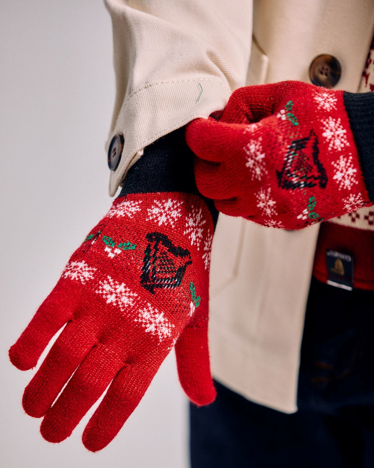 Red gloves with snowflake and bell pattern worn by a person in a beige coat.