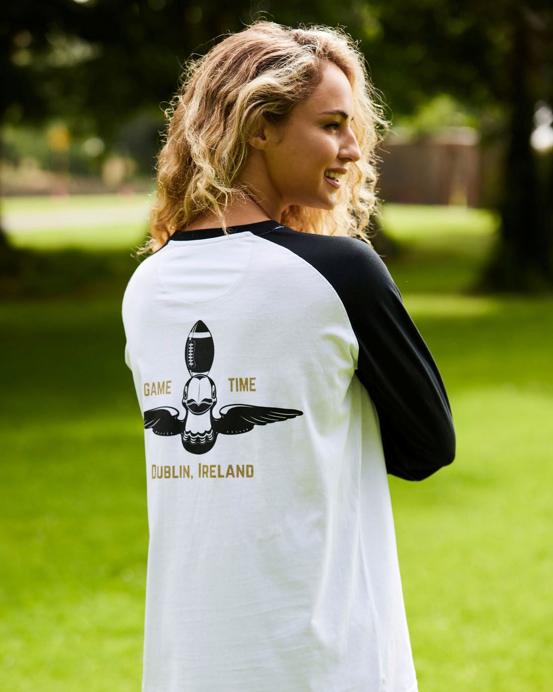 Woman wearing a white and black baseball shirt with a graphic and text on the back, standing outdoors.