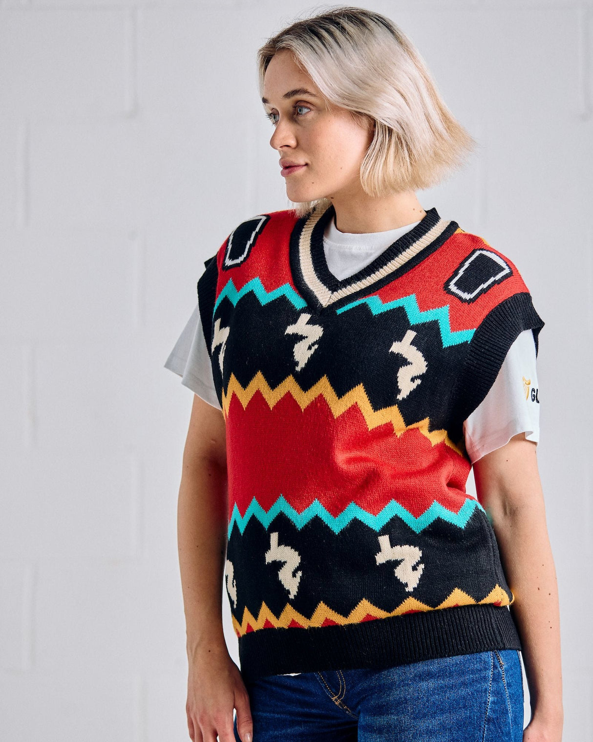 Person wearing a colorful patterned vest over a white shirt with blue jeans against a light gray background