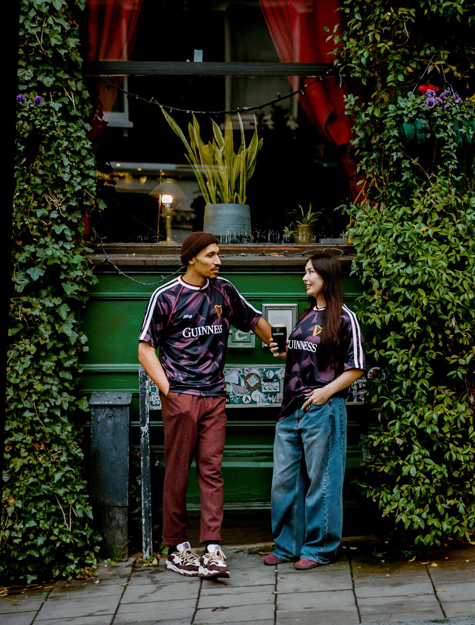 Two people wearing matching Guinness-themed outfits standing in front of a green vehicle with ivy-covered walls.