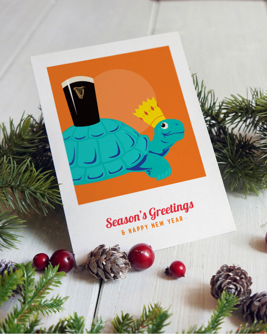 Greeting card with a turtle wearing a crown and a pint of Guinness, surrounded by Christmas decorations on a white surface.