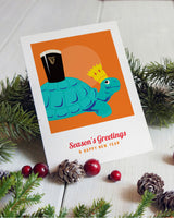 Greeting card with a turtle wearing a crown and a pint of Guinness, surrounded by Christmas decorations on a white surface.