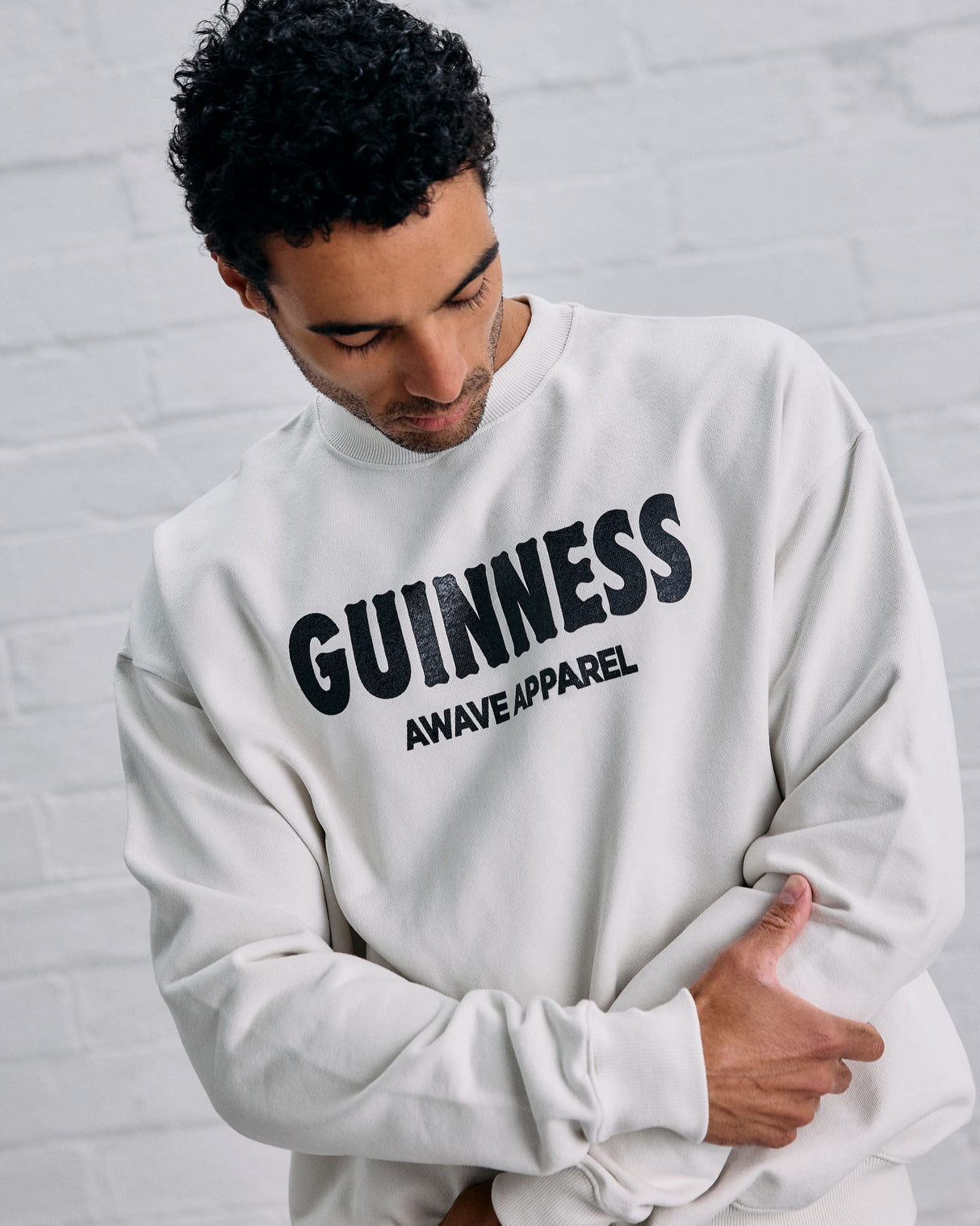Man wearing a white sweatshirt with 'Guinness Awave Apparel' text against a gray brick wall.