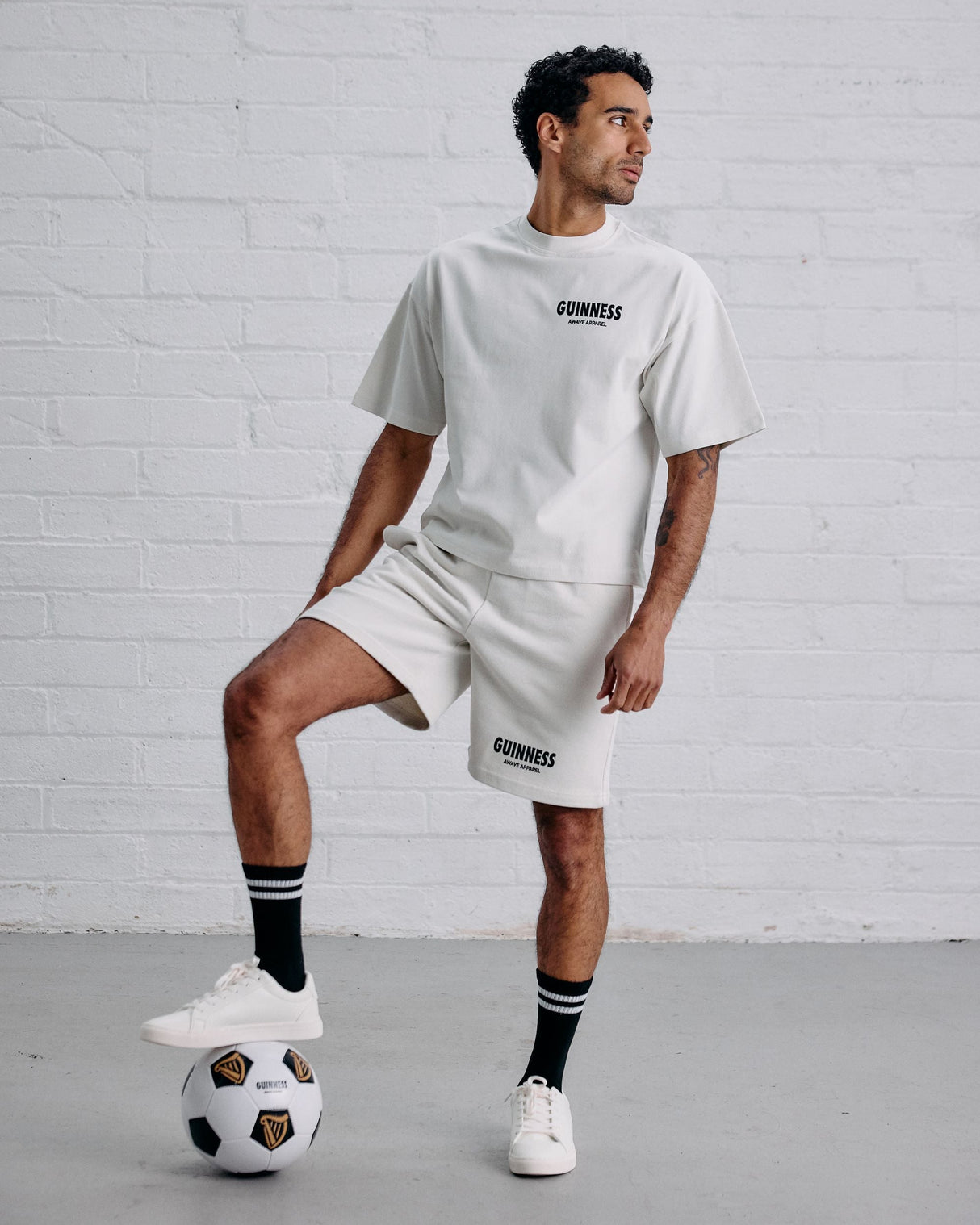 Man wearing a white 'Guinness' t-shirt and shorts with a soccer ball on a gray floor against a white brick wall.