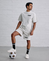 Man wearing a white 'Guinness' t-shirt and shorts with a soccer ball on a gray floor against a white brick wall.