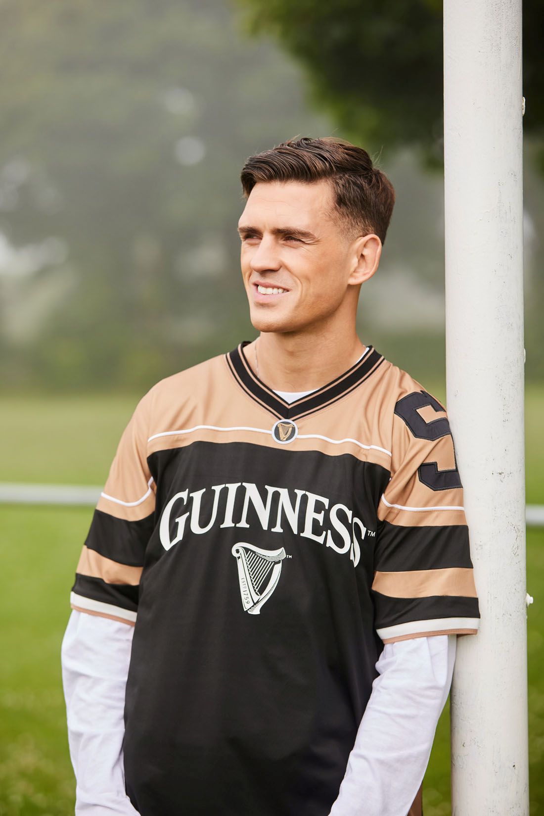 Man wearing a Guinness jersey leaning against a pole outdoors.
