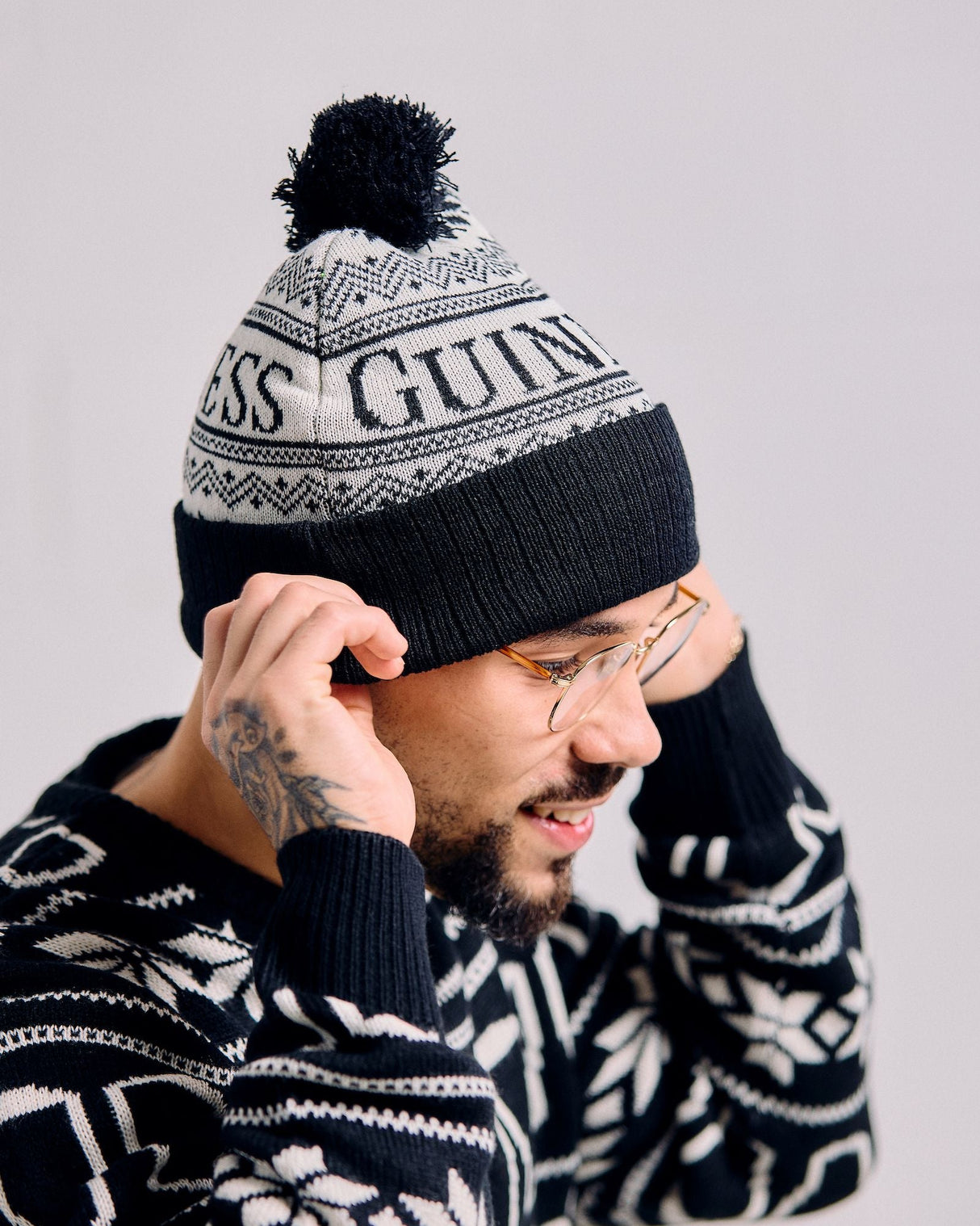 Person wearing a black and white knit beanie with pom-pom and a patterned sweater on a light gray background