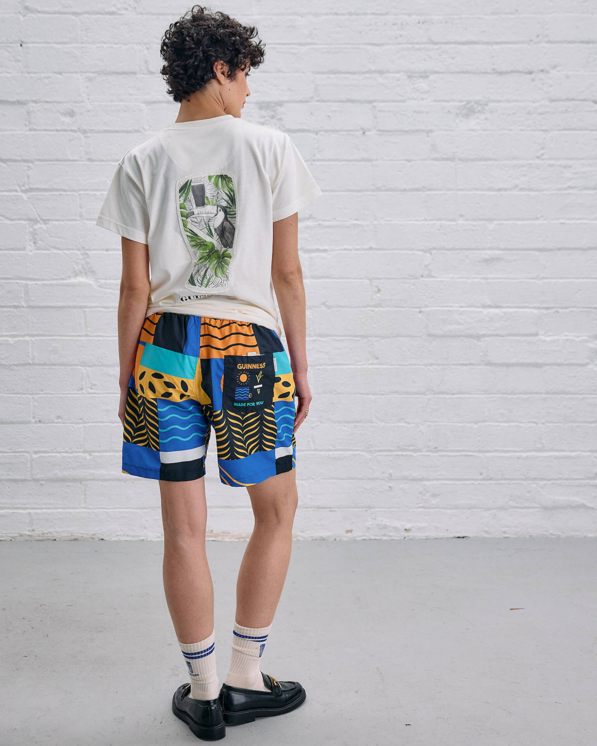 Person wearing a white t-shirt with a graphic design and colorful shorts against a white brick wall.
