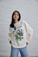 Woman wearing a white sweatshirt with a graphic design in front of a white brick wall.