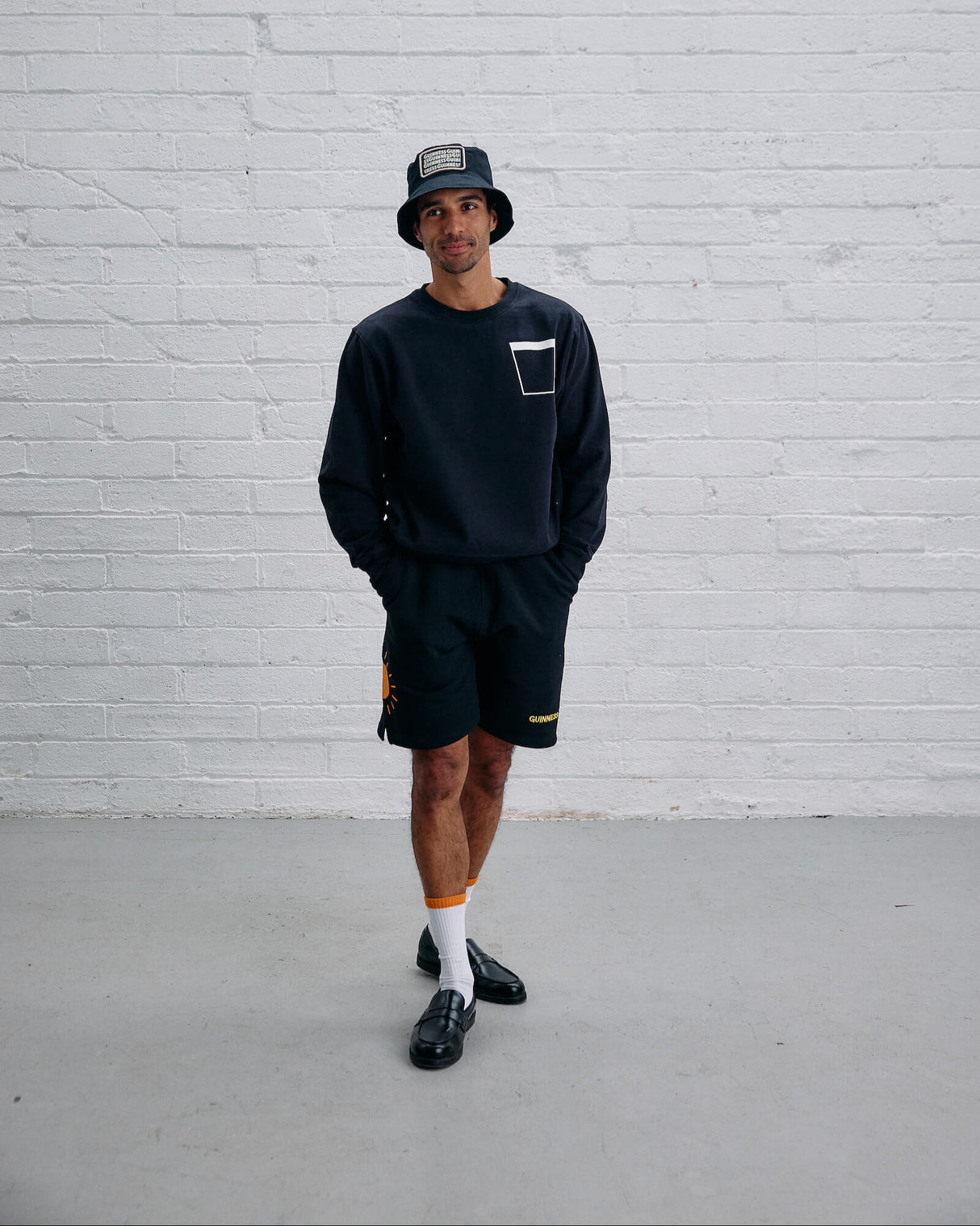 Man wearing a black sweatshirt, shorts, and hat against a white brick wall.
