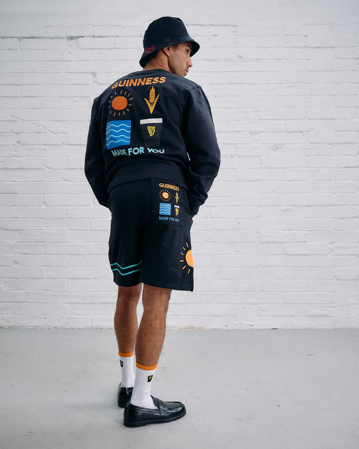 Person wearing a black outfit with colorful patches against a white brick wall.