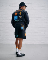 Person wearing a black outfit with colorful patches against a white brick wall.