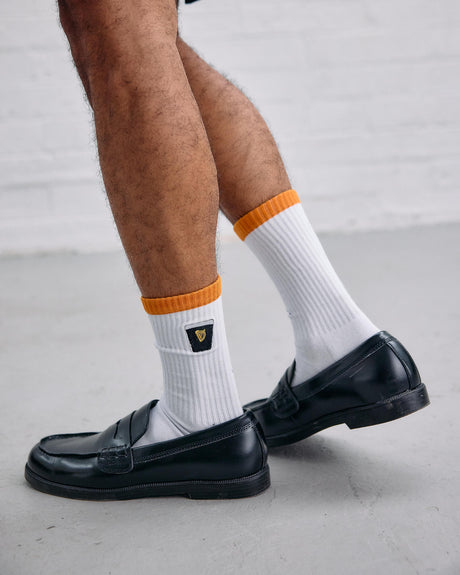 Person wearing white socks with orange accents and black loafers on a light gray background