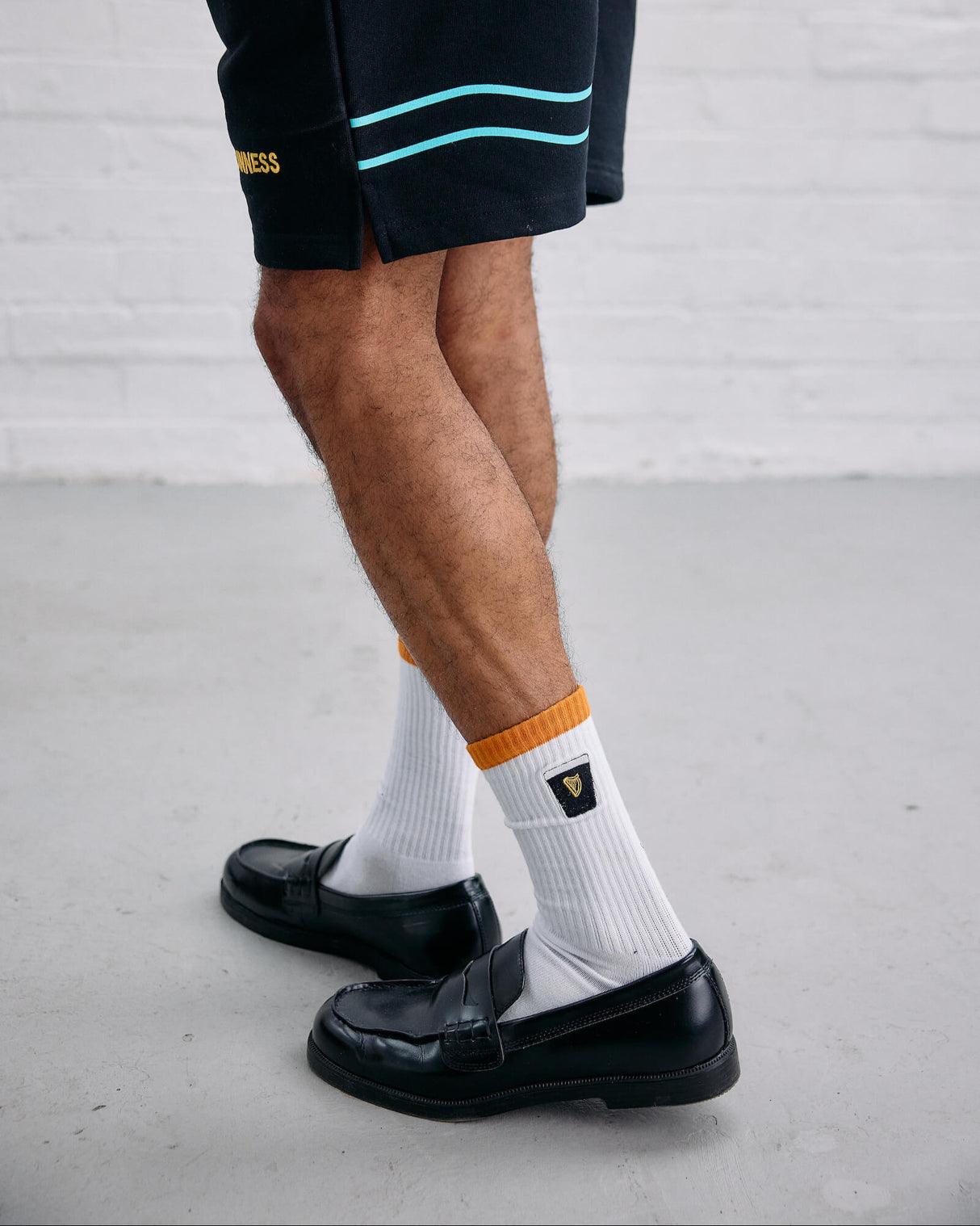 Person wearing black shorts with a brand logo, white socks with an orange label, and black shoes on a light gray floor.