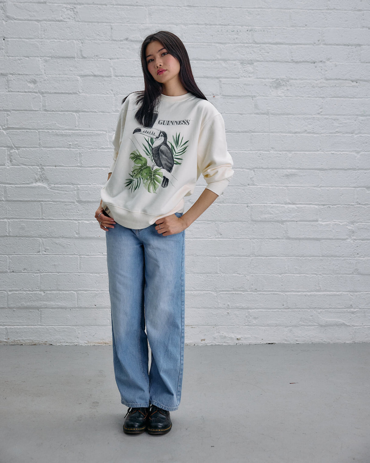 Person wearing a graphic sweatshirt and jeans against a white brick wall.