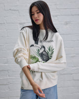 Woman wearing a white sweatshirt with a graphic design against a light gray brick wall.