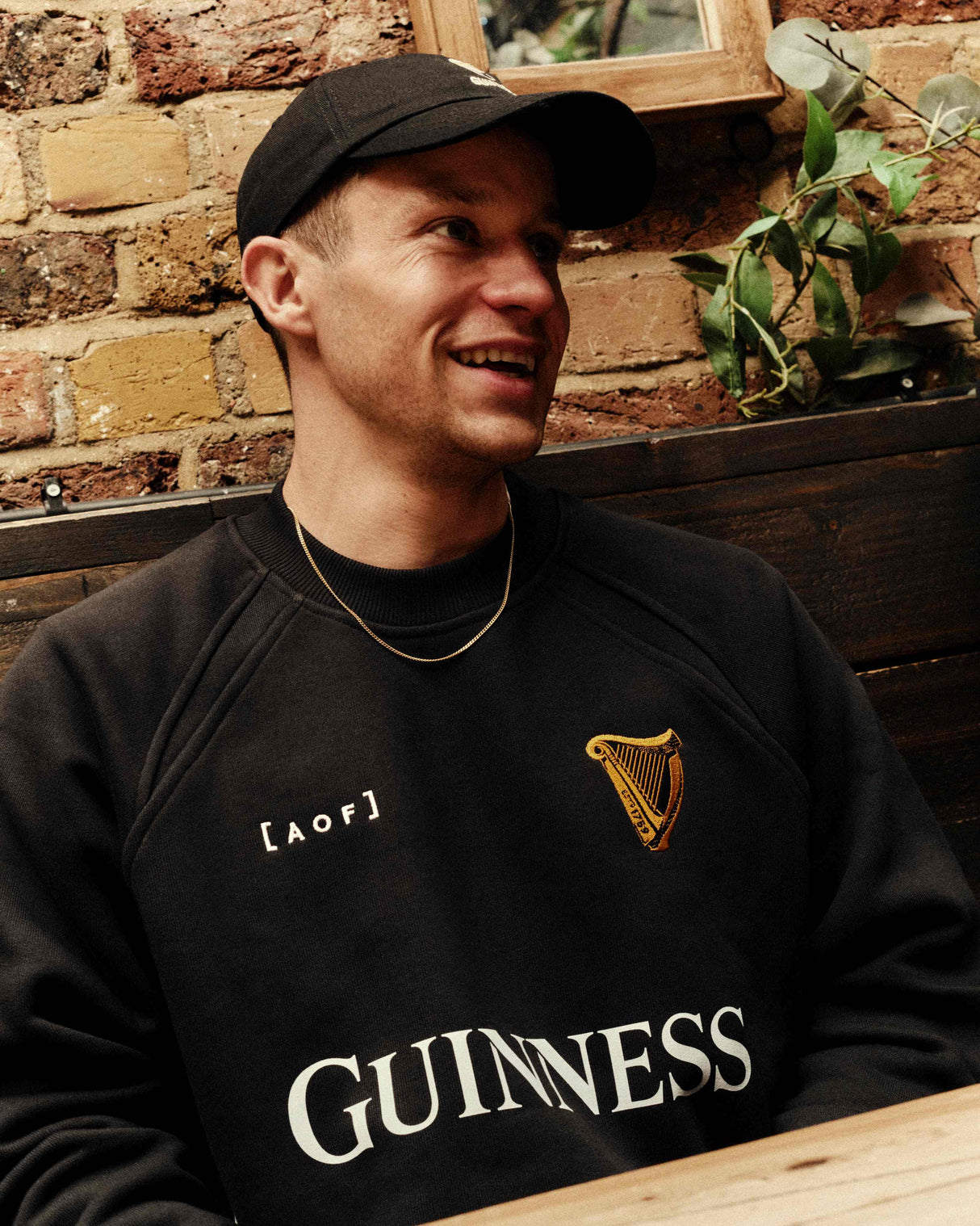 Man wearing a black Guinness sweatshirt with a logo, sitting against a brick wall.