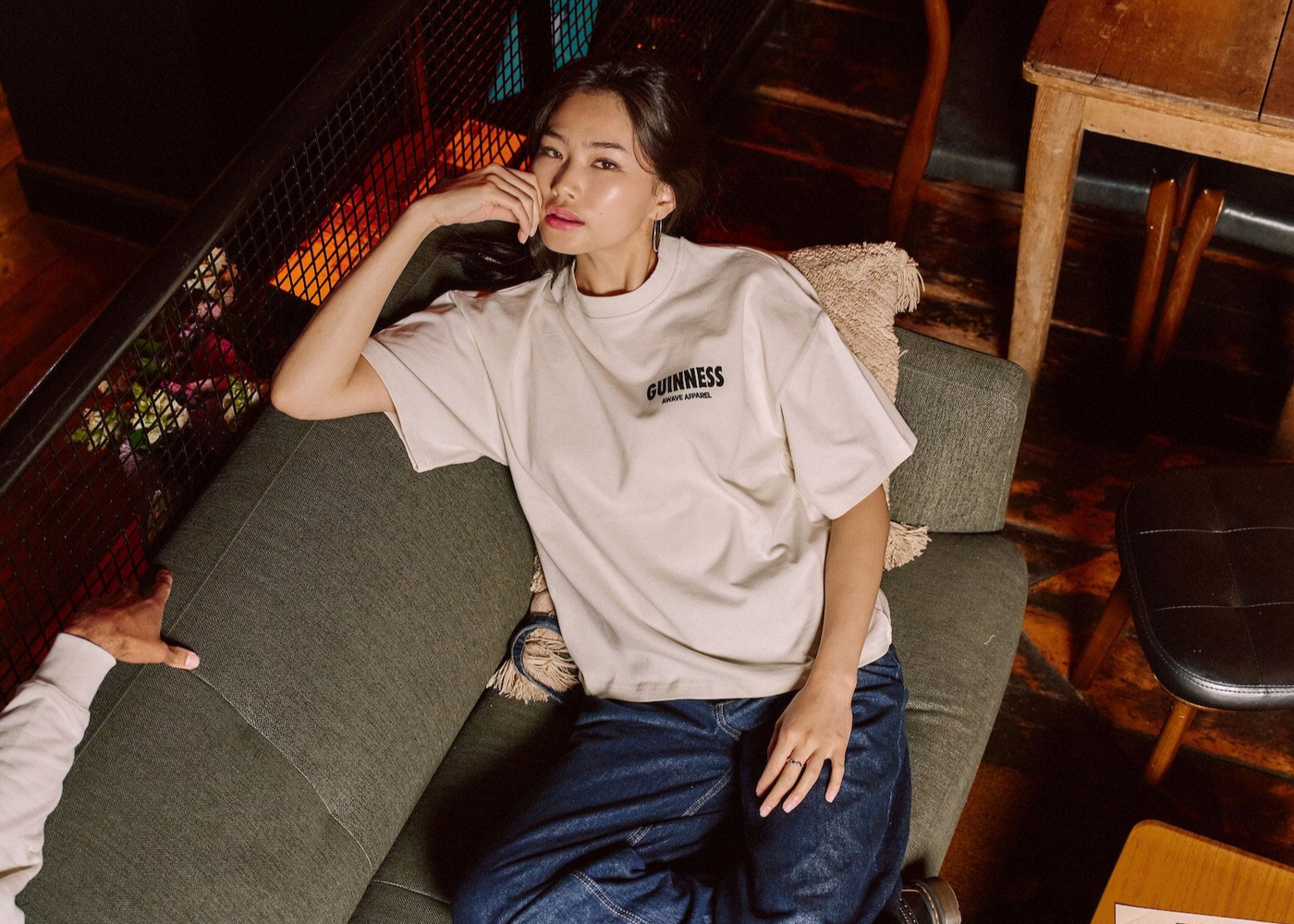 Woman sitting on a couch wearing a white t-shirt with Guinness logo and blue jeans in an indoor setting.