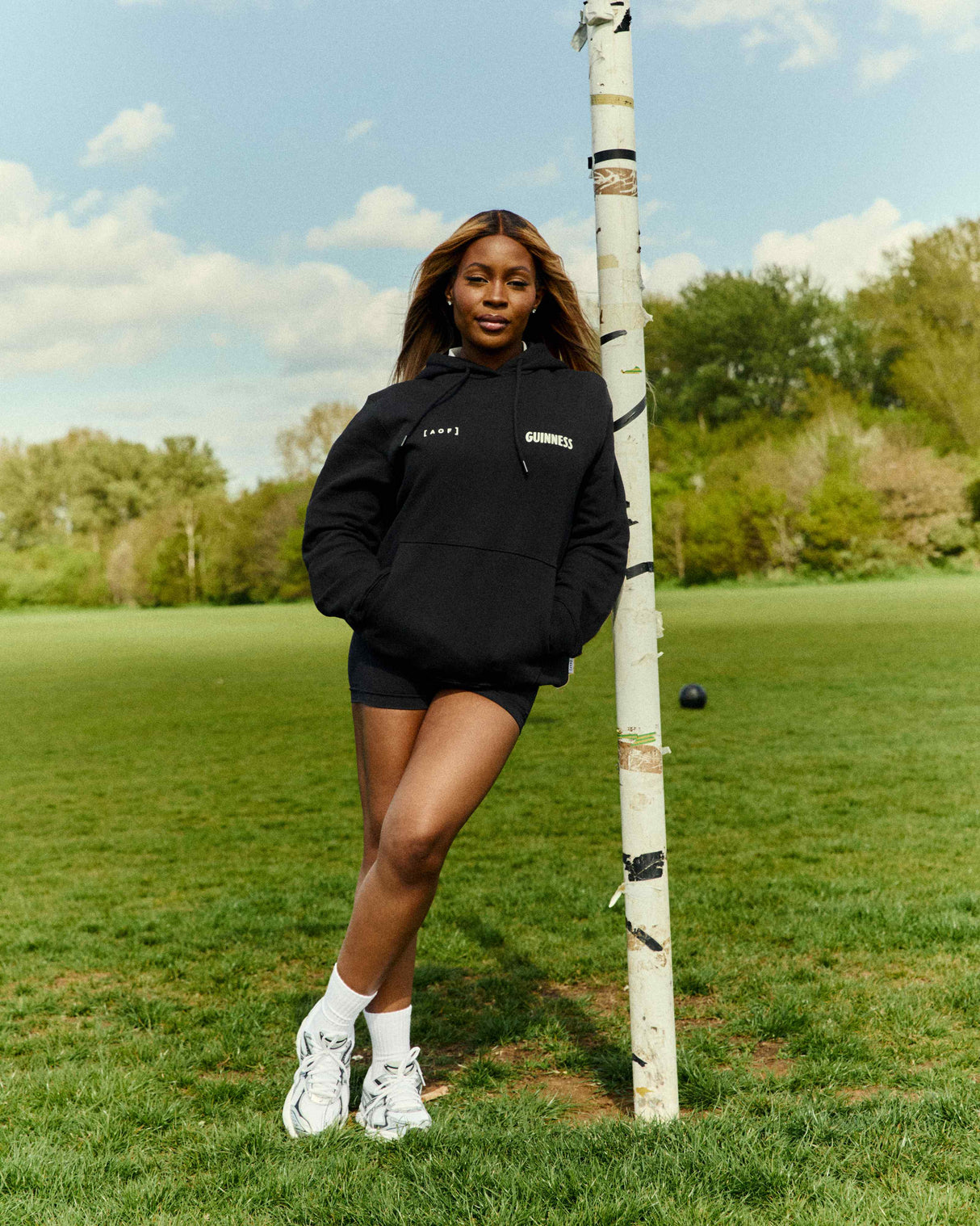 Person wearing a black hoodie and shorts standing next to a birch tree on a grassy field.