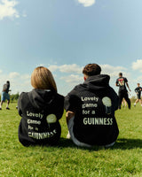Two people sitting on a grassy field wearing black hoodies with text, looking at a sports game.