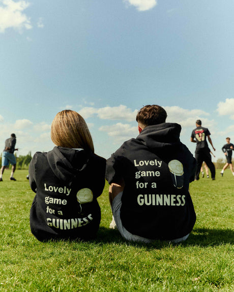 Two people sitting on a grassy field wearing black hoodies with text, looking at a sports game.