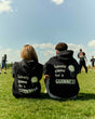 Two people sitting on a grassy field wearing black hoodies with text, looking at a sports game.