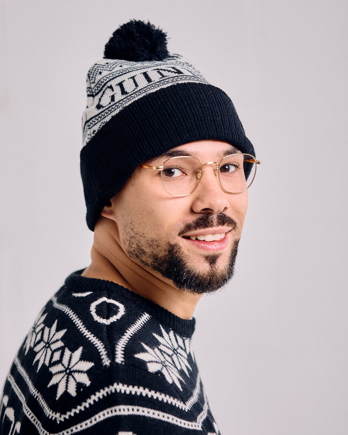 Person wearing a black knit beanie with a pom-pom and a matching sweater with white patterns on a light gray background