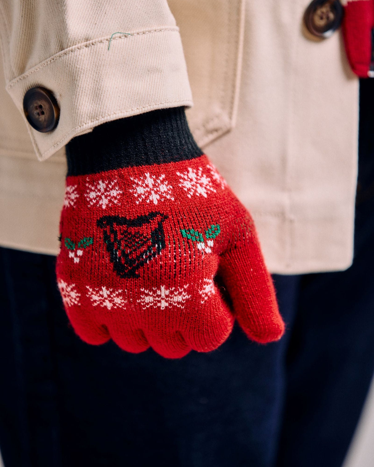 Red knitted gloves with festive design held by a person wearing a beige coat.