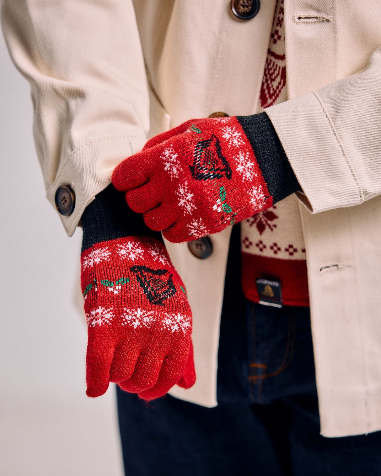 Red gloves with festive patterns worn by a person in a beige coat and dark pants.