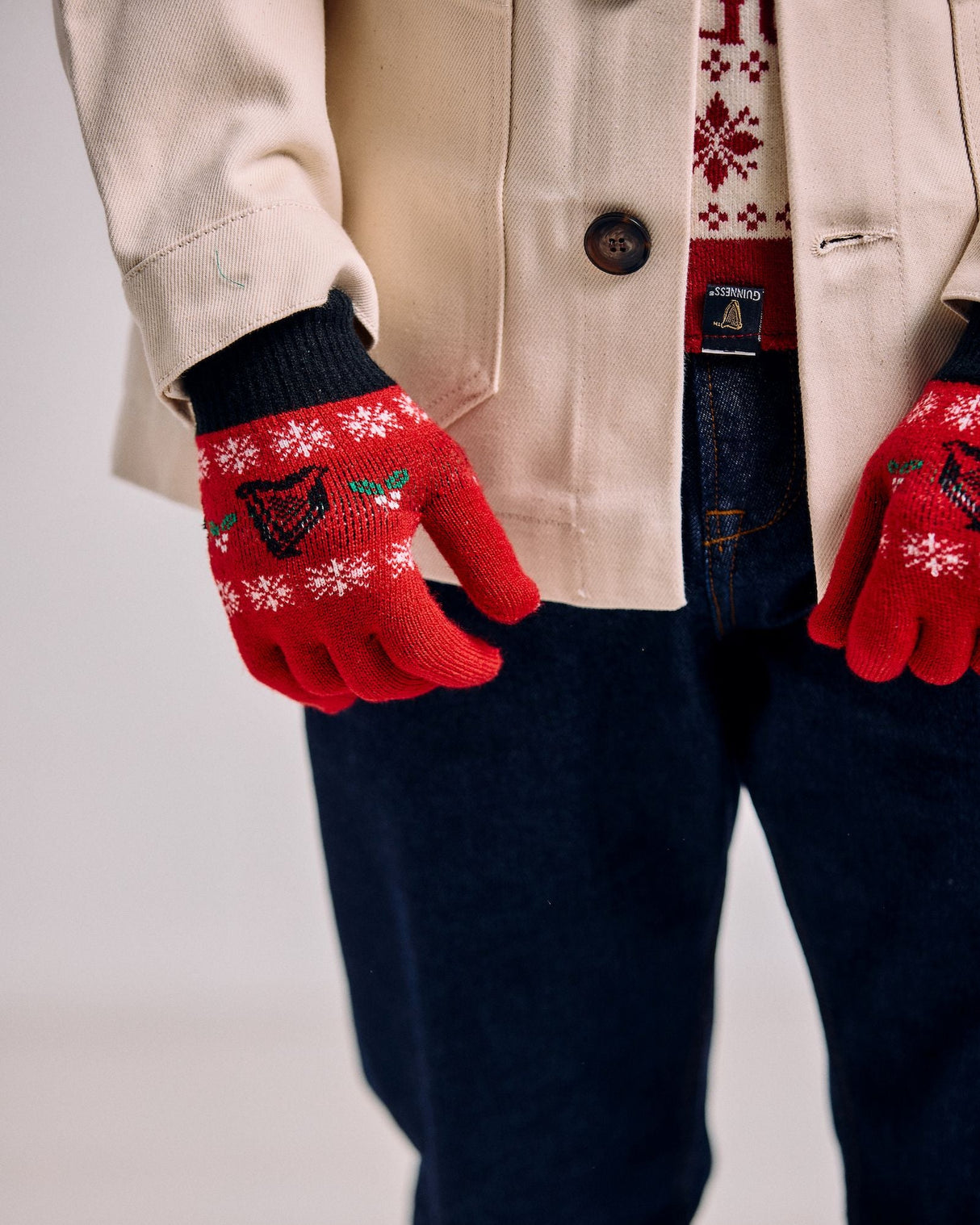 Person wearing red gloves with snowflake patterns, a beige coat, and dark pants on a light background