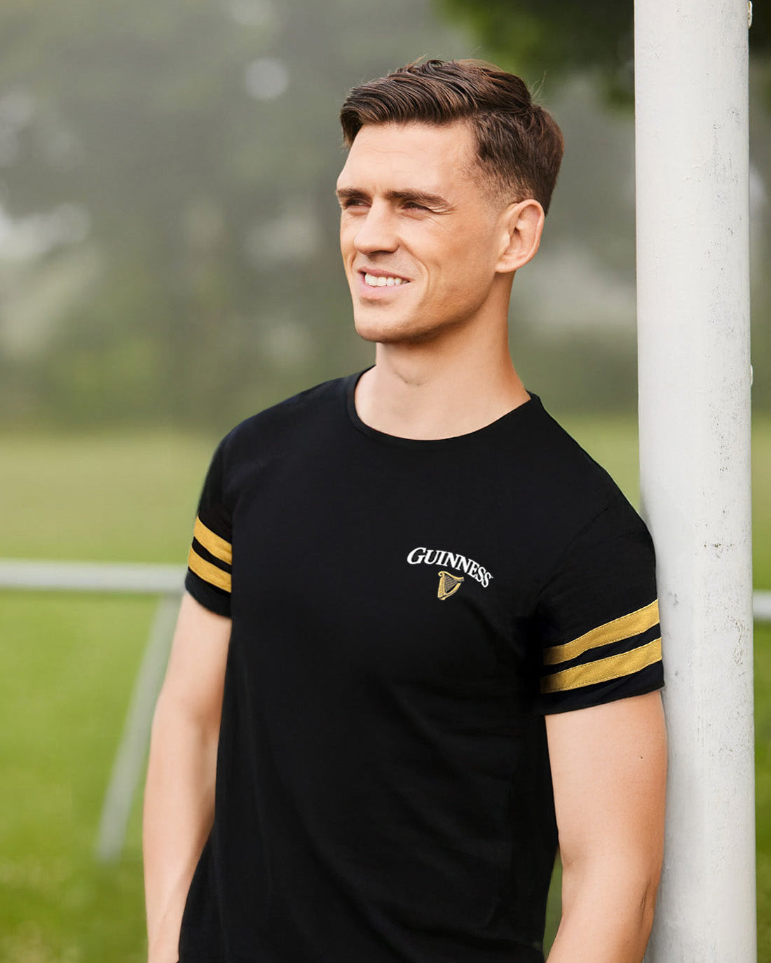 Man wearing a black t-shirt with yellow stripes and a visible brand logo, standing outdoors.