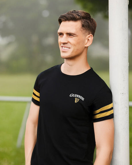 Man wearing a black t-shirt with yellow stripes and a visible brand logo, standing outdoors.