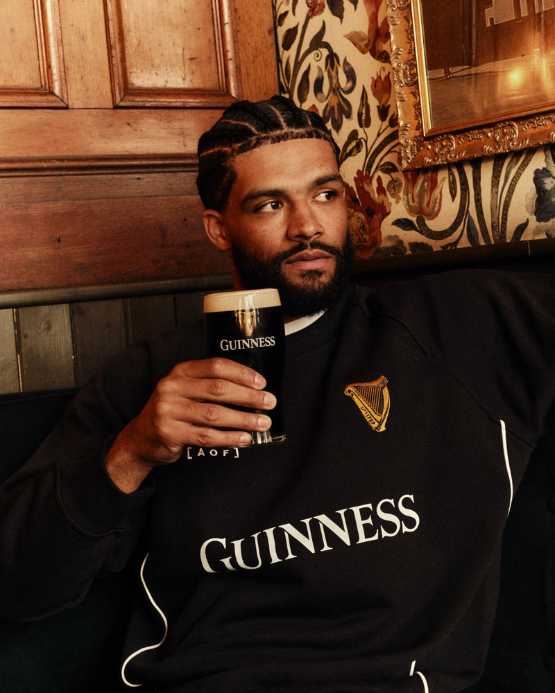 Man wearing a black Guinness sweatshirt holding a pint of Guinness in a pub setting.