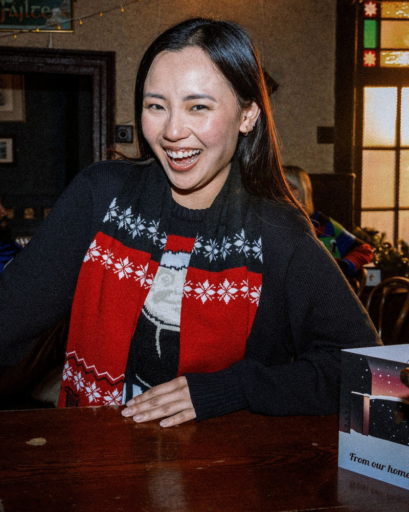 Woman wearing a festive sweater with a red and black pattern, sitting at a table in a warm-lit room.