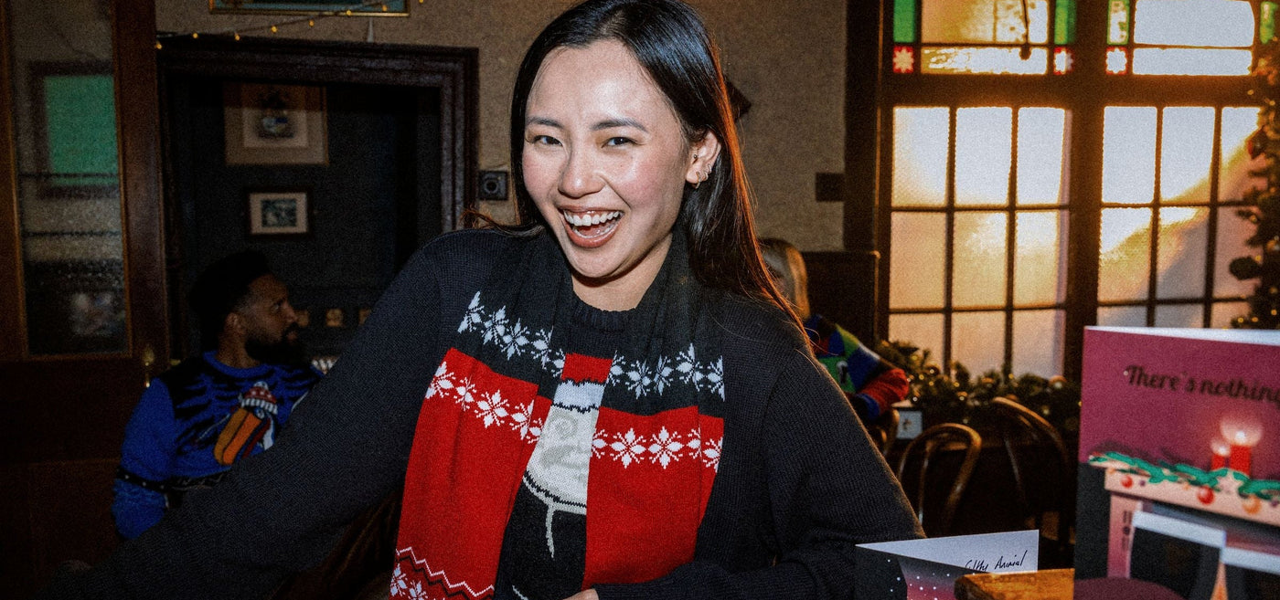Woman wearing a festive sweater with a red and black pattern, sitting at a table in a warm-lit room.