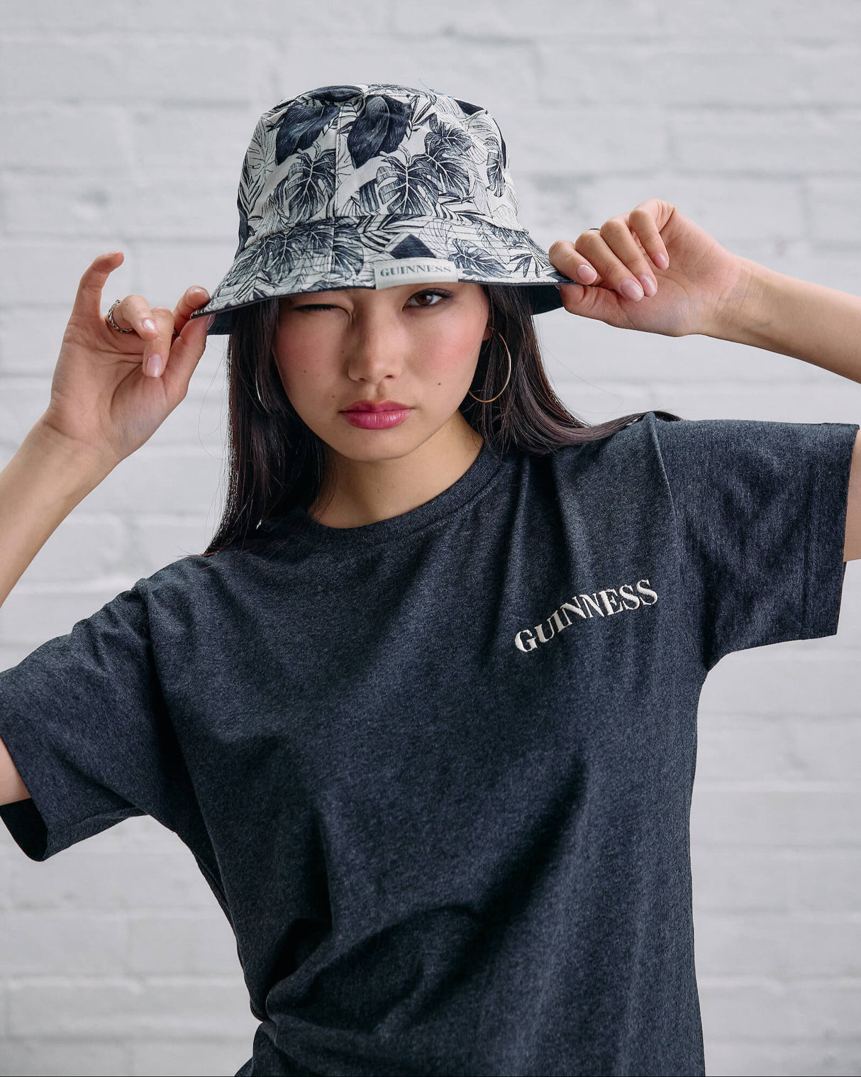 Person wearing a patterned bucket hat and dark t-shirt with 'Guinness' logo against a white brick wall.