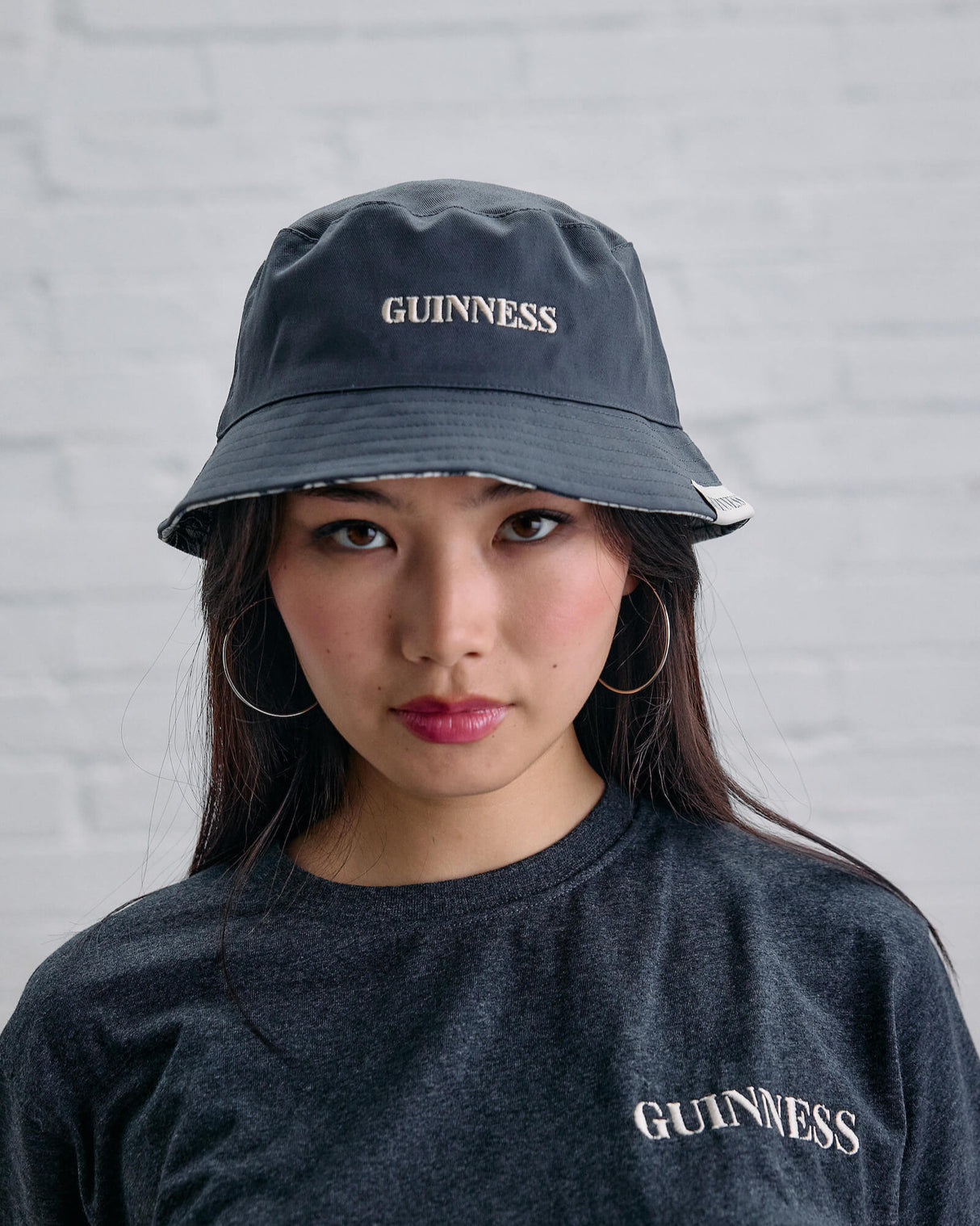 Person wearing a black 'Guinness' bucket hat and shirt against a gray brick wall.