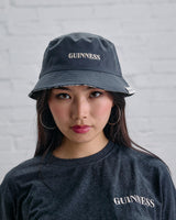 Person wearing a black 'Guinness' bucket hat and shirt against a gray brick wall.