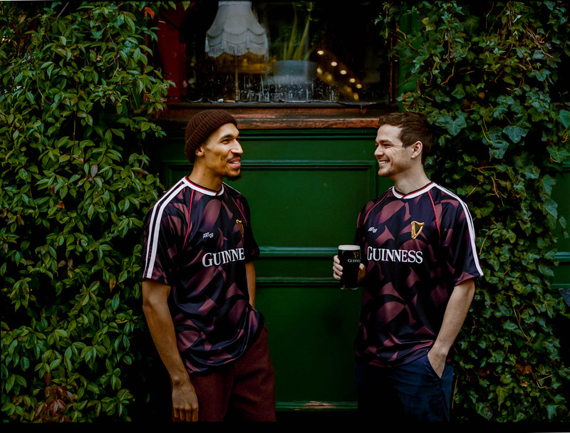 Two men wearing Guinness-themed jerseys standing in front of a green wall with plants.